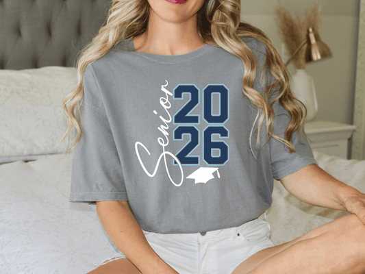 A woman wearing a grey t-shirt with the text "Graduation 26" printed on it, sitting on a bed.