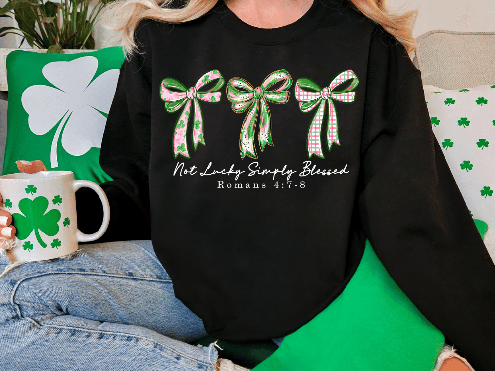 A person wearing a black sweatshirt with a graphic design of three green bows and the text "Not Lucky Simply Blessed" printed on it. The person is sitting on a couch with a shamrock-themed mug and a green pillow.
