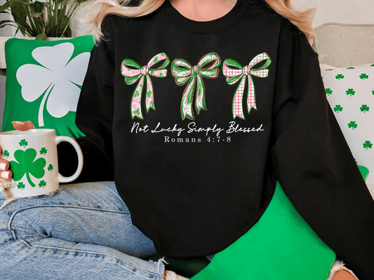 A person wearing a black sweatshirt with a graphic design of three green bows and the text "Not Lucky Simply Blessed" printed on it. The person is sitting on a couch with a shamrock-themed mug and a green pillow.