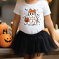 A young girl wearing a white t-shirt with a ghost graphic and a black tulle skirt stands in front of a collection of carved jack-o-lanterns.