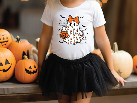 A young girl wearing a white t-shirt with a ghost graphic and a black tulle skirt stands in front of a collection of carved jack-o-lanterns.