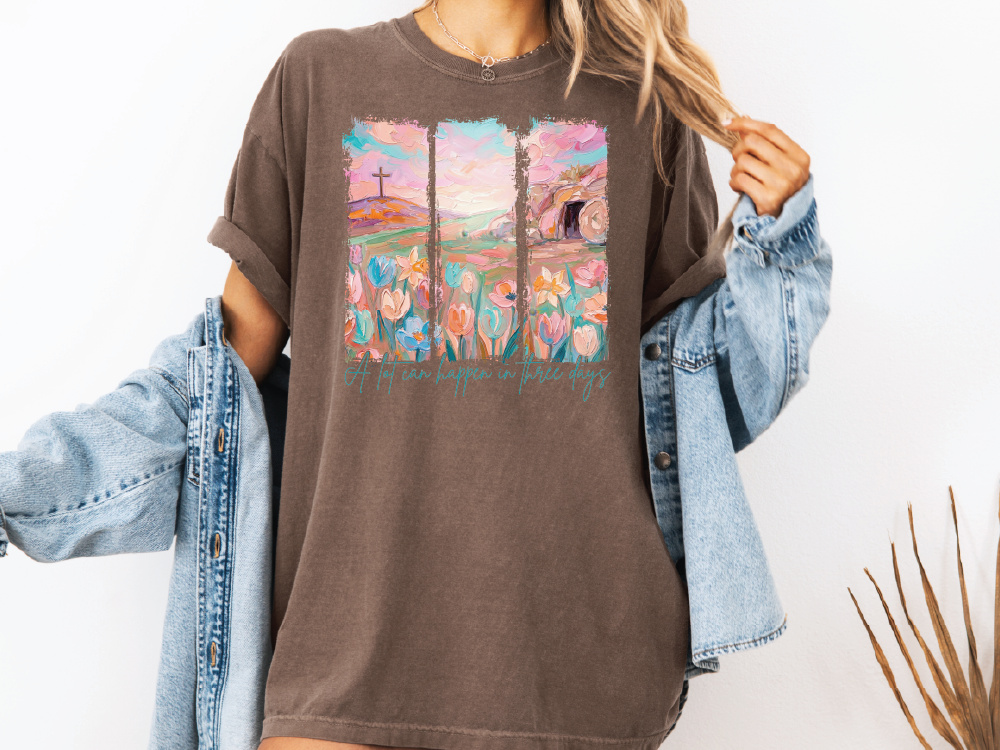 A woman wearing a brown t-shirt with a colorful graphic design and denim jacket, standing against a white background.