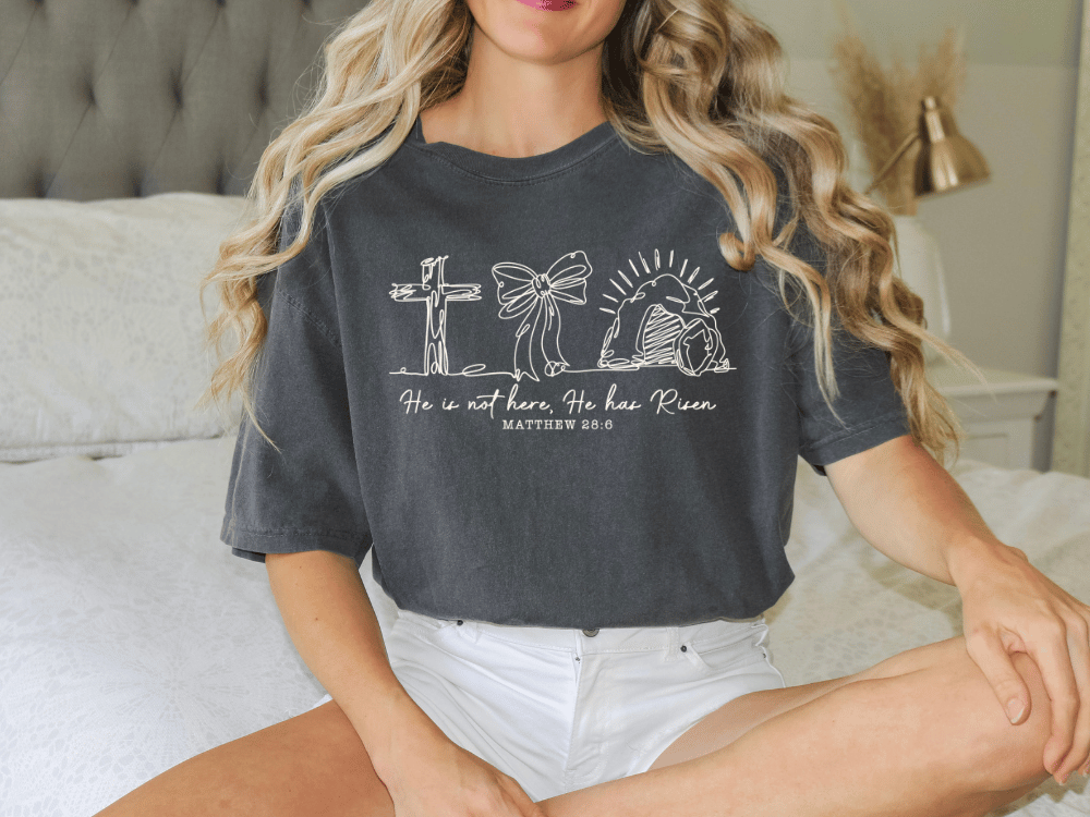 A woman wearing a gray t-shirt with a graphic design and text printed on it, sitting on a bed.