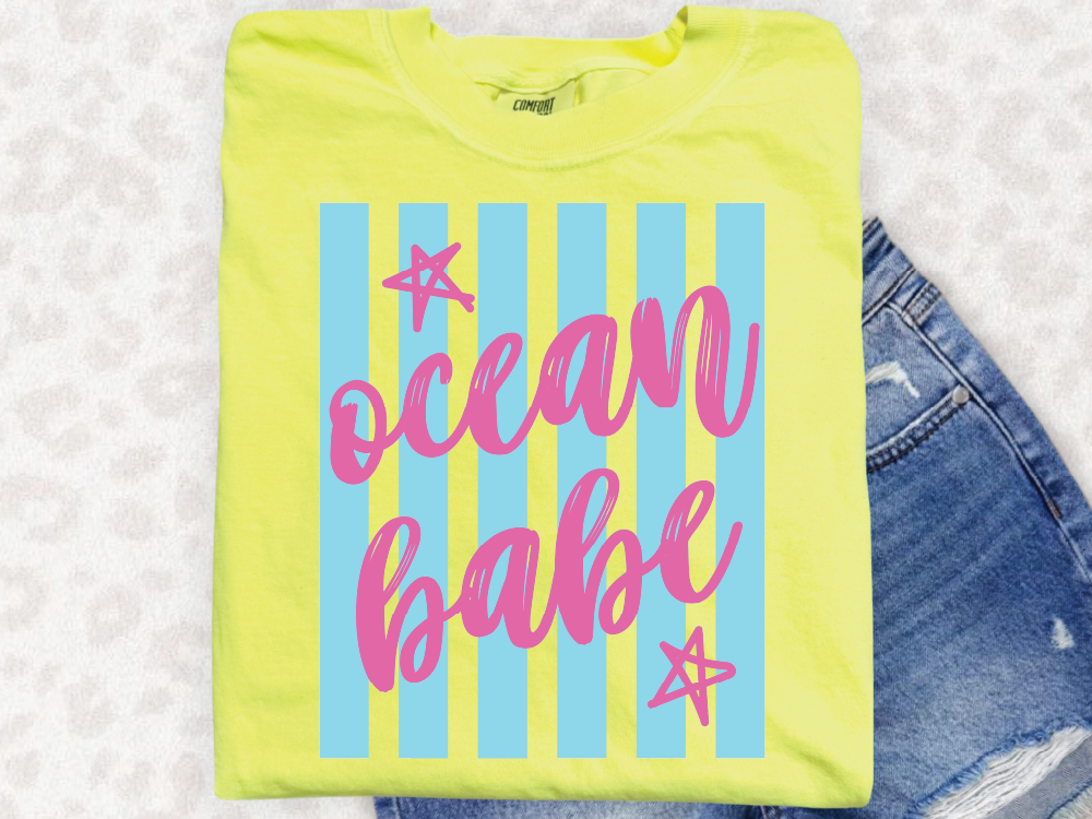 A yellow t-shirt with the text "ocean babe" printed on it, along with pink and blue striped designs.