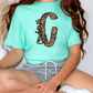 A person wearing a turquoise t-shirt with a leopard print design and gray shorts is sitting on the floor.