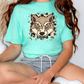 A person wearing a turquoise t-shirt with a graphic of a wolf's face and a white skirt is sitting cross-legged on the floor.