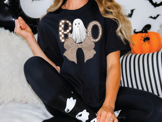 A person wearing a black t-shirt with a graphic of a ghost and a bow, sitting on a couch with Halloween decorations in the background.