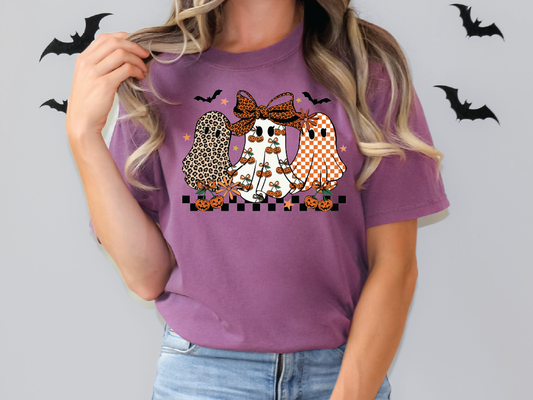 A woman wearing a purple t-shirt with a graphic design of three ghostly figures, including a leopard-print ghost, a checkered ghost, and a pumpkin-wearing ghost.