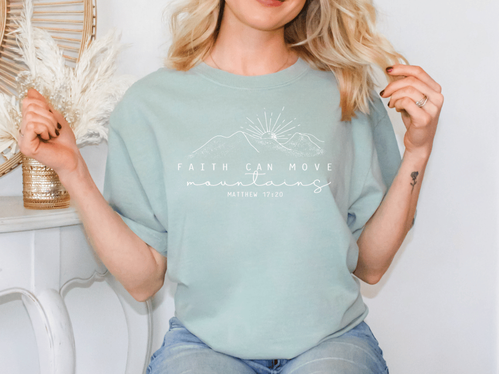 A woman wearing a light blue t-shirt with a mountain and sun graphic, and the text "Faith can move mountains" printed on it.