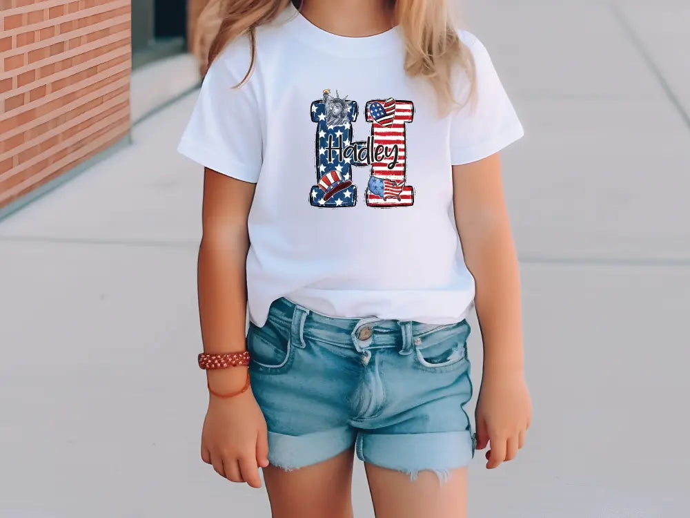 a little girl standing on a sidewalk wearing a t - shirt with the letter h