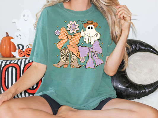 A woman wearing a green t-shirt with a graphic of a ghost, bow, and boots, sitting on a couch with Halloween decorations in the background.
