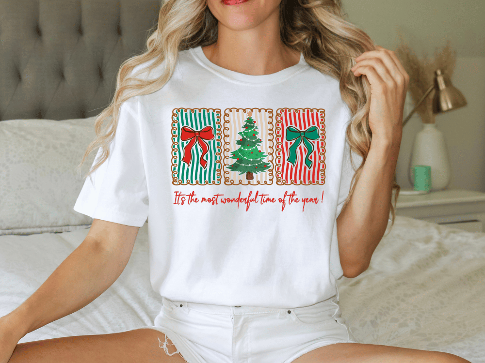 A woman wearing a white t-shirt with a Christmas-themed graphic design, including a Christmas tree and bows, sitting on a bed.