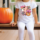 A young girl wearing a white t-shirt with a Halloween-themed graphic sits on a wooden bench next to a large orange pumpkin.