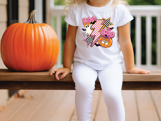 A young girl wearing a white t-shirt with a Halloween-themed graphic sits on a wooden bench next to a large orange pumpkin.