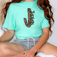 A person wearing a turquoise t-shirt with a leopard print design and gray shorts is sitting on the floor.