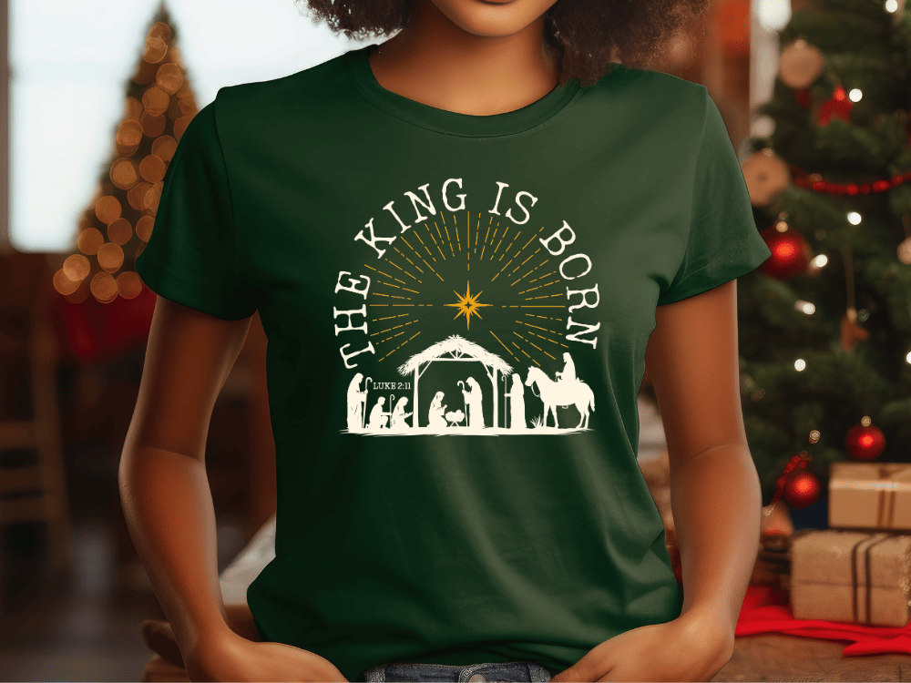 A woman wearing a green t-shirt with a graphic design depicting a nativity scene and the text "The King is Born" printed on it.