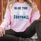 A person wearing a pink sweatshirt with the text "BLUE TIDE FOOTBALL" printed on it, sitting on a blanket surrounded by pumpkins.