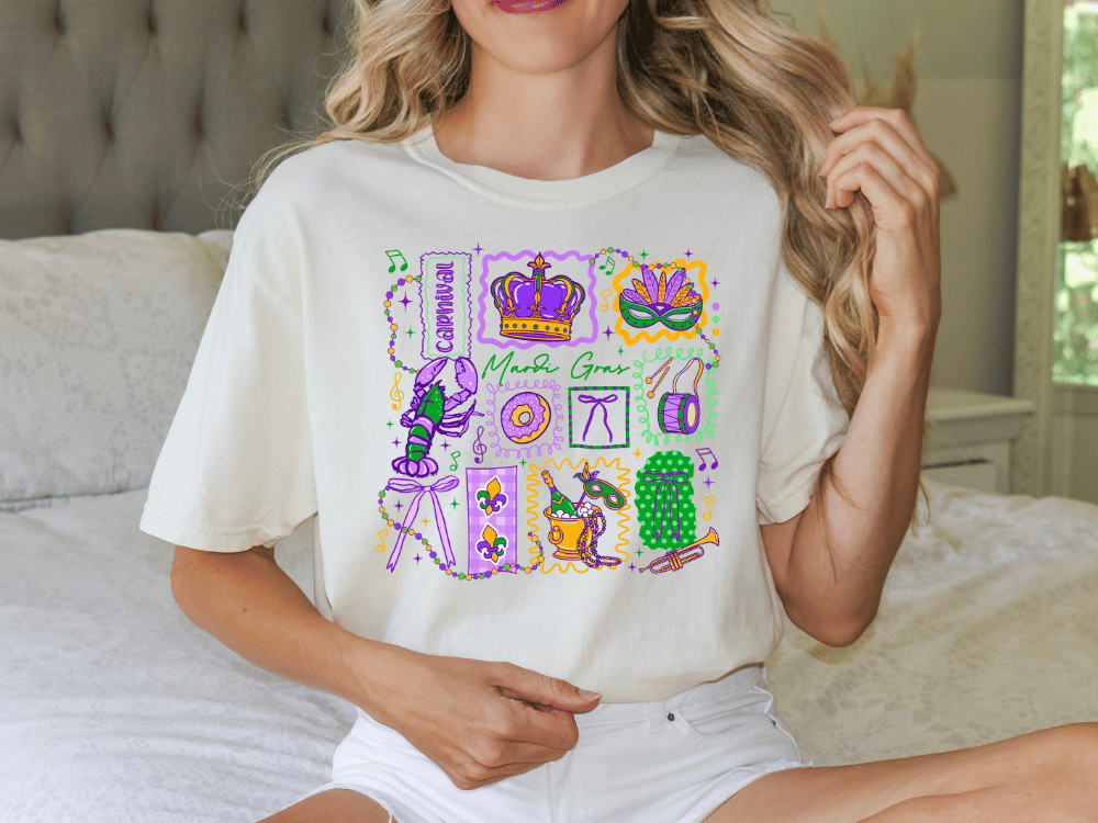 A woman wearing a white t-shirt with a colorful graphic design featuring various Mardi Gras-related elements such as a crown, mask, and beads.
