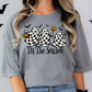 A woman wearing a gray t-shirt with a graphic design featuring three ghostly figures holding pumpkins and the text "Tis The Season" against a background of bats.