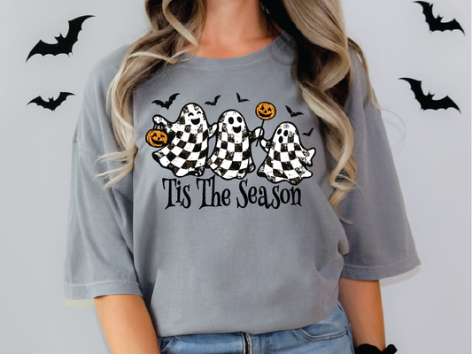 A woman wearing a gray t-shirt with a graphic design featuring three ghostly figures holding pumpkins and the text "Tis The Season" against a background of bats.