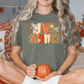 A woman sitting on a bed with a pumpkin-themed pillow and a mug in her hands, wearing a green t-shirt with a Thanksgiving-themed design.