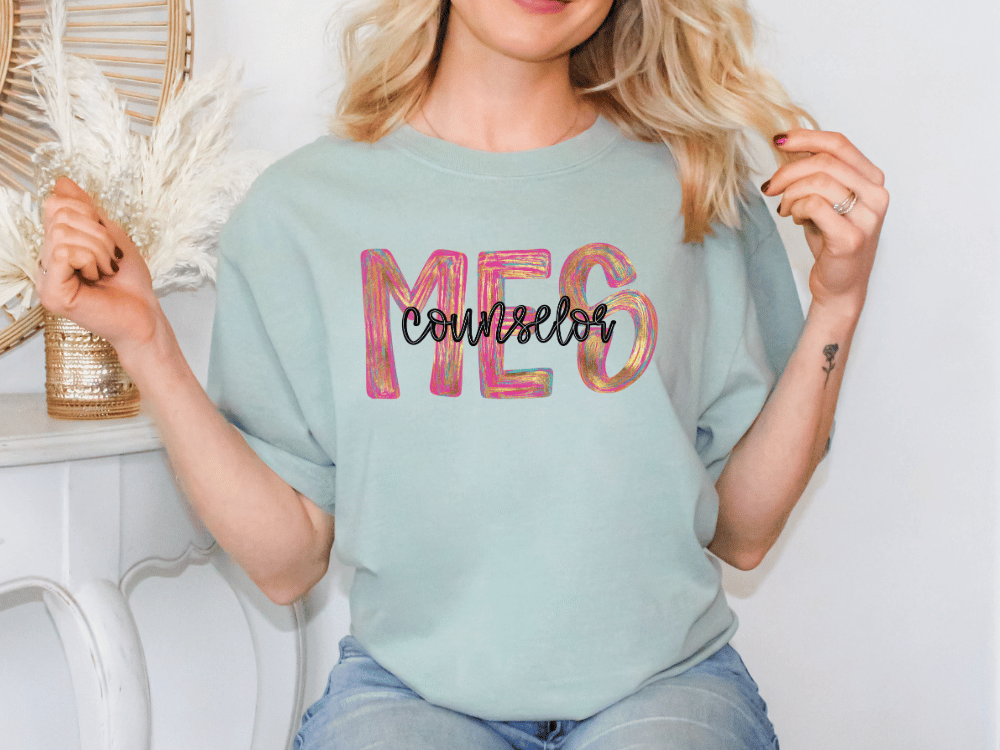 A woman wearing a light blue t-shirt with the word "Counselor" printed on it, sitting in front of a white wall with a decorative item behind her.