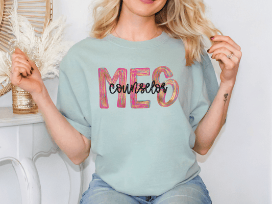 A woman wearing a light blue t-shirt with the word "Counselor" printed on it, sitting in front of a white wall with a decorative item behind her.