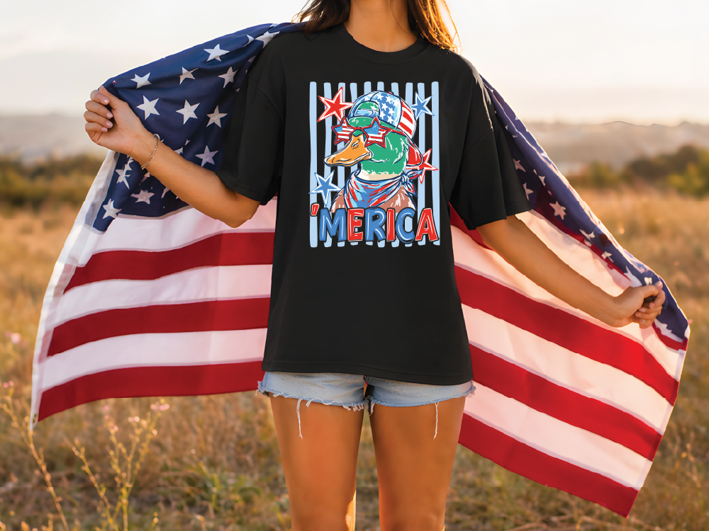 A woman stands in a field holding an American flag, wearing a black t-shirt with a graphic design featuring a duck wearing sunglasses and the word "MERICA".