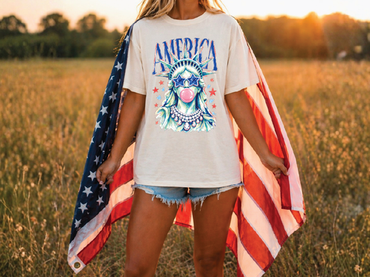A woman stands in a field, wearing a white t-shirt with an American flag design and holding the flag behind her back.