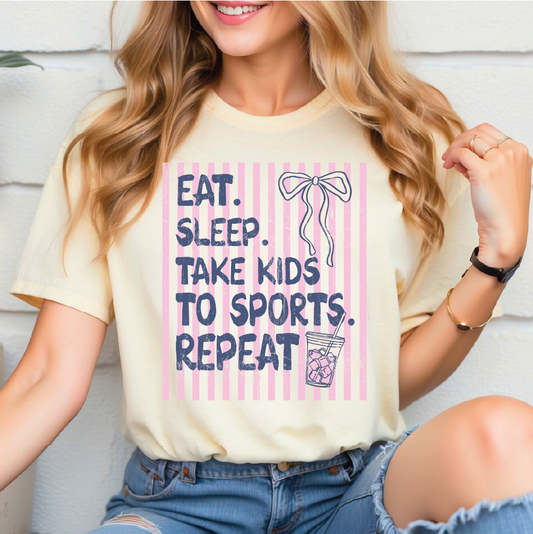 A woman wearing a yellow t-shirt with a graphic design that says "EAT. SLEEP. TAKE KIDS TO SPORTS. REPEAT" along with a bow and a cup of soda.