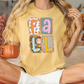 A woman wearing a yellow t-shirt with the word "FALL" printed on it, sitting on a bed with a pumpkin-shaped mug in her hand.