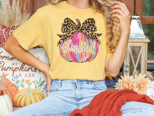 A woman wearing a yellow t-shirt with a pumpkin graphic and the word "Thankful" is sitting on a bench surrounded by fall decorations.