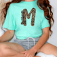 A person wearing a mint green t-shirt with a leopard print design and gray shorts is sitting on the floor.