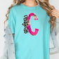 A woman wearing a turquoise t-shirt with a large pink and black letter 'C' printed on it, along with a denim jacket.