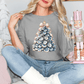A woman is sitting on a couch, wearing a gray t-shirt with a Christmas tree design, and holding a nutcracker.