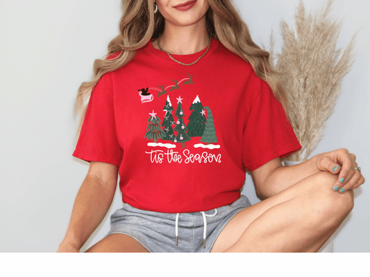 A woman wearing a red t-shirt with a Christmas tree design and the text "Tis the Season" is sitting against a white background.