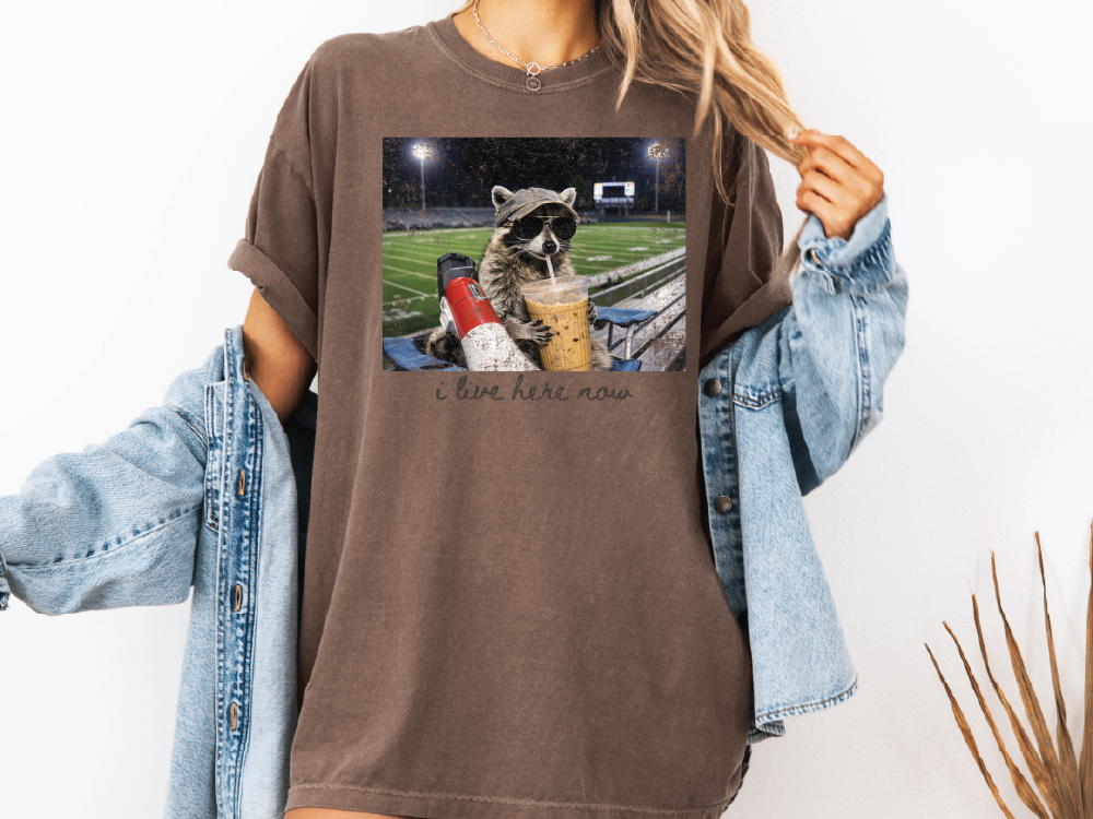 A person wearing a brown t-shirt with a graphic of a raccoon sitting on a bleachers and holding a drink, along with the text "I love here now".