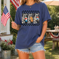 A woman wearing a blue t-shirt with a graphic of raccoons holding hot dogs, standing in front of a patriotic setting with an American flag and a picnic table.