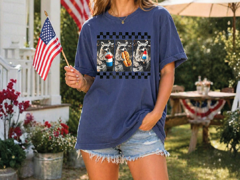 A woman wearing a blue t-shirt with a graphic of raccoons holding hot dogs, standing in front of a patriotic setting with an American flag and a picnic table.