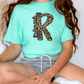 A person wearing a turquoise t-shirt with a leopard print design and gray shorts is sitting on the floor.
