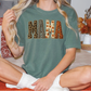 A woman wearing a green t-shirt with the word "MAMA" printed on it, sitting on a bed with autumn-themed pillows and a vase of flowers in the background.