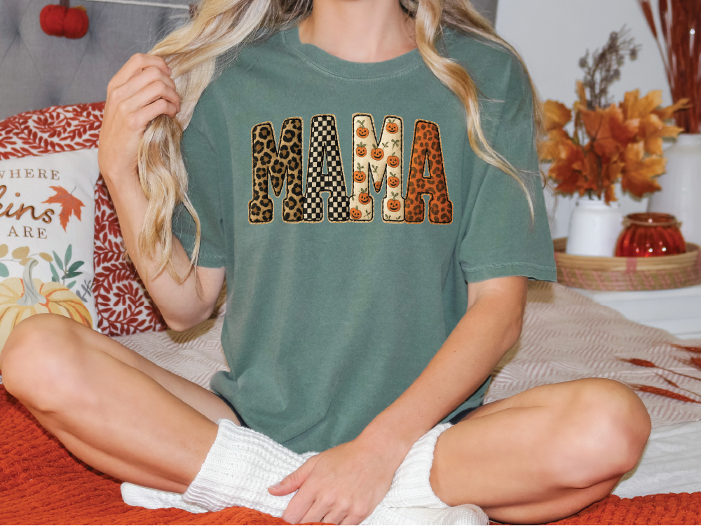 A woman wearing a green t-shirt with the word "MAMA" printed on it, sitting on a bed with autumn-themed pillows and a vase of flowers in the background.