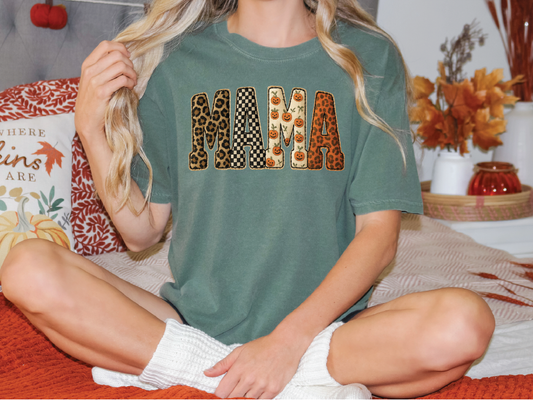 A woman wearing a green t-shirt with the word "MAMA" printed on it, sitting on a bed with autumn-themed pillows and a vase of flowers in the background.
