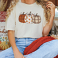 A woman wearing a white t-shirt with a pumpkin graphic and blue jeans is sitting on a bench with a red blanket and a pumpkin-themed pillow.