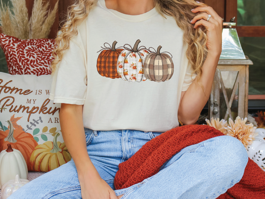 A woman wearing a white t-shirt with a pumpkin graphic and blue jeans is sitting on a bench with a red blanket and a pumpkin-themed pillow.