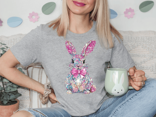 A woman wearing a gray t-shirt with a colorful, glittery bunny graphic is sitting on a couch, holding a green mug.