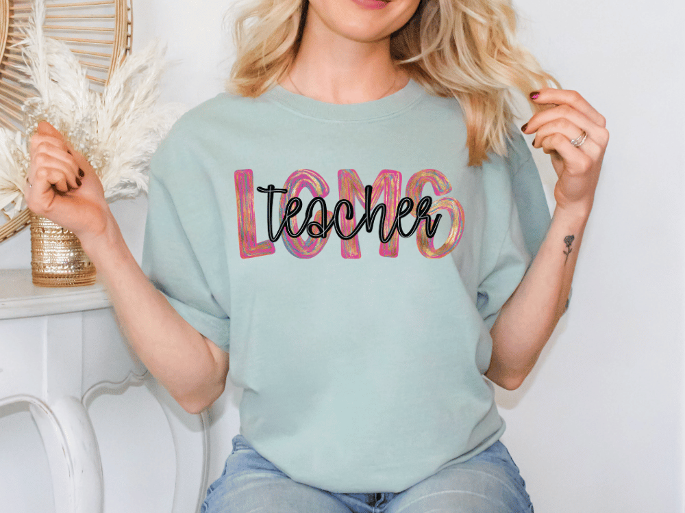 A woman wearing a light blue t-shirt with the word "teacher" printed on it, smiling and holding her hair.