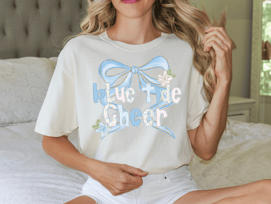 A woman wearing a white t-shirt with a blue bow and the text "Blue Tide Cheer" is sitting on a bed.
