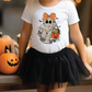 A young girl wearing a white t-shirt with a ghost graphic and a black tulle skirt stands in front of a display of Halloween pumpkins.