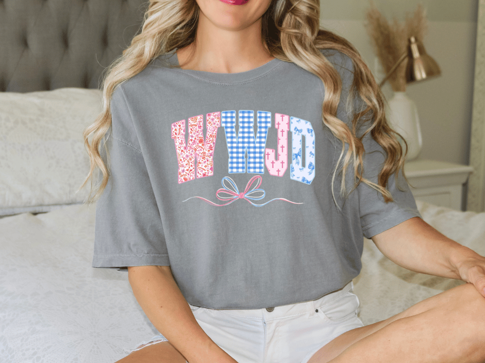 A woman wearing a gray t-shirt with the word "WHJD" printed on it, sitting on a bed.